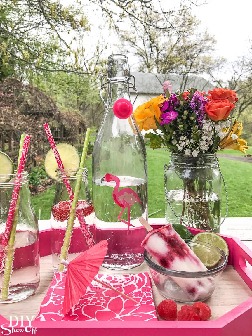 Summer drink station with raspberry lime popsicle in a bowl, fresh limes, glass bottles with fruit-infused water, and a vase of colorful flowers on a patio table