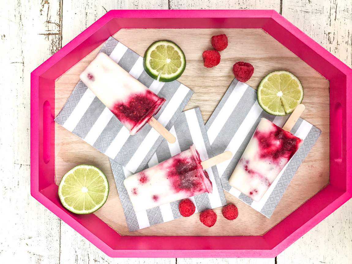 Overhead view of striped raspberry lime essential oil popsicles with fresh lime slices and raspberries on a pink tray